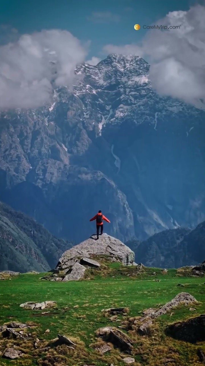 A traveller standing on a rock in a green Himalayan meadow below snow peaks